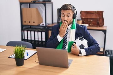 Handsome hispanic man watching football game at the office covering mouth with hand, shocked and...