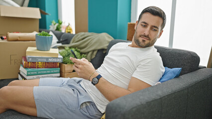 Obraz premium Young hispanic man using smartphone sitting on sofa at new home