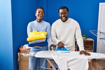 Young african american couple ironing clothes at laundry room smiling and laughing hard out loud because funny crazy joke.