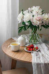 A bouquet of white peonies in a glass vase, chamomile tea, strawberries on a white round table in a cozy living room