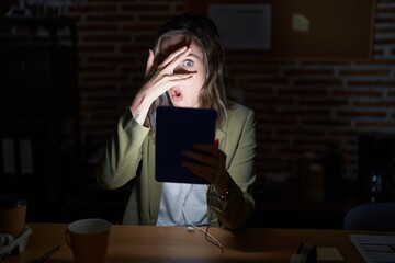 Blonde caucasian woman working at the office at night peeking in shock covering face and eyes with hand, looking through fingers with embarrassed expression.