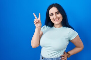 Fototapeta premium Young modern girl with blue hair standing over blue background smiling looking to the camera showing fingers doing victory sign. number two.