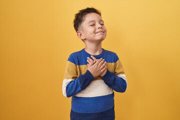 Little hispanic boy standing over yellow background smiling with hands on chest, eyes closed with...