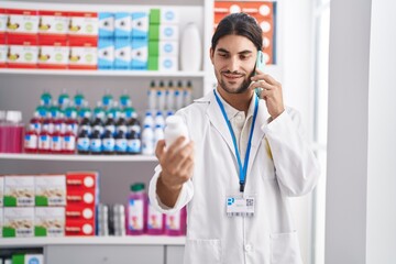 Young hispanic man pharmacist holding pills bottle talking on smartphone at pharmacy