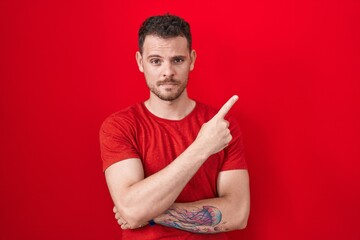 Young hispanic man standing over red background pointing with hand finger to the side showing advertisement, serious and calm face