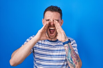 Young hispanic man standing over blue background shouting angry out loud with hands over mouth