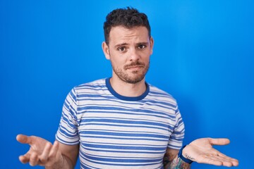 Young hispanic man standing over blue background clueless and confused with open arms, no idea concept.
