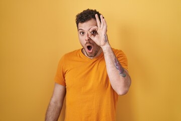 Young hispanic man standing over yellow background doing ok gesture shocked with surprised face, eye looking through fingers. unbelieving expression.