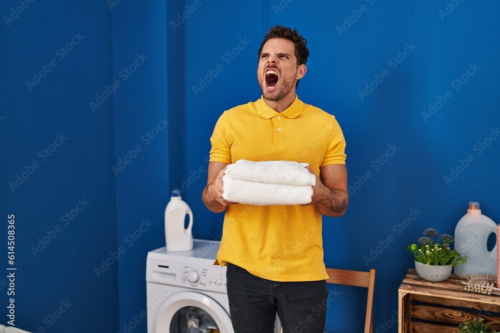 Poster young hispanic man holding clean towels at laundry room angry and mad screaming frustrated and furio