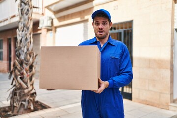 Young hispanic man delivering box in shock face, looking skeptical and sarcastic, surprised with...
