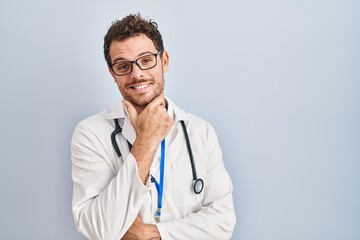 Young hispanic man wearing doctor uniform and stethoscope looking confident at the camera smiling with crossed arms and hand raised on chin. thinking positive.