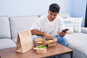 Young hispanic man eating take away food using smartphone at home