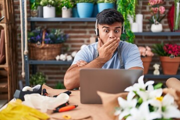 Hispanic young man working at florist shop doing video call covering mouth with hand, shocked and afraid for mistake. surprised expression