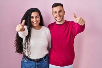 Young hispanic couple standing over pink background approving doing positive gesture with hand, thumbs up smiling and happy for success. winner gesture.