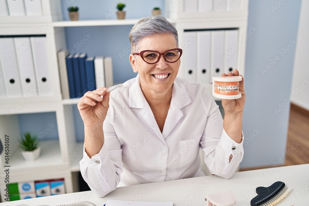 Sticker Middle age woman wearing dentist uniform holding brackets and aligner at clinic