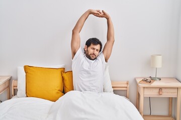 Young hispanic man waking up sitting on bed at bedroom