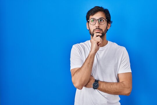 Handsome latin man standing over blue background with hand on chin thinking about question, pensive expression. smiling with thoughtful face. doubt concept.