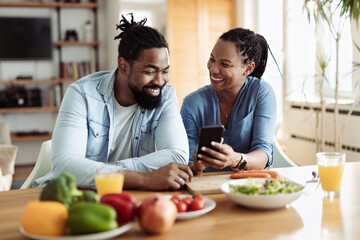 Fototapeta na wymiar Happy African American couple using a phone while having breakfast in the morning. Focus is on woman
