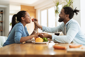 Happy black man feeding his girlfriend with tomato during lunch in dining room. Focus is on woman