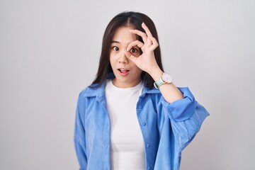 Young chinese woman standing over white background doing ok gesture shocked with surprised face, eye looking through fingers. unbelieving expression.