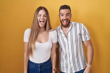 Young couple standing over yellow background sticking tongue out happy with funny expression. emotion concept.