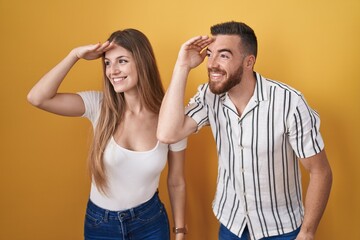 Young couple standing over yellow background very happy and smiling looking far away with hand over head. searching concept.