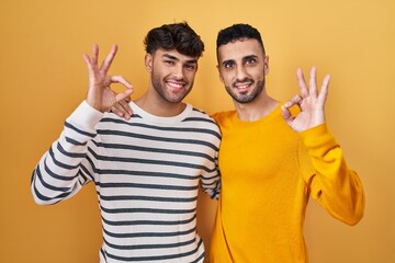 Fototapeta premium Young hispanic gay couple standing over yellow background smiling positive doing ok sign with hand and fingers. successful expression.