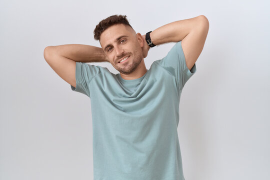 Hispanic man with beard standing over white background relaxing and stretching, arms and hands behind head and neck smiling happy