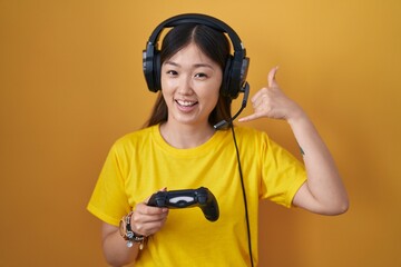 Chinese young woman playing video game holding controller smiling doing phone gesture with hand and fingers like talking on the telephone. communicating concepts.