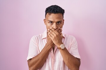 Young hispanic man standing over pink background shocked covering mouth with hands for mistake....