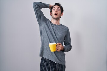 Young hispanic man wearing pajama drinking a cup of coffee crazy and scared with hands on head,...