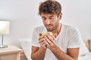 Young man smelling cup of coffee sitting on bed at bedroom
