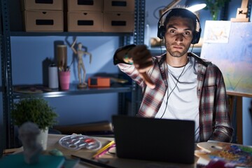 Young hispanic man sitting at art studio with laptop late at night looking unhappy and angry...