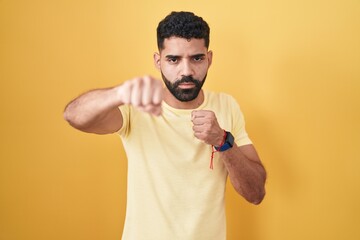 Hispanic man with beard standing over yellow background punching fist to fight, aggressive and...