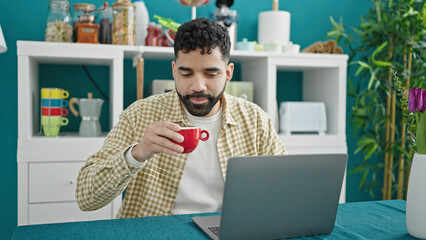Young hispanic man using laptop drinking coffee at dinning room