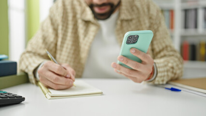 Young hispanic man student using smartphone writing notes at library university