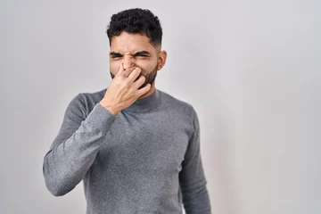 Foto op Canvas Hispanic man with beard standing over white background smelling something stinky and disgusting, intolerable smell, holding breath with fingers on nose. bad smell © Krakenimages.com