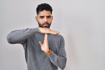 Hispanic man with beard standing over white background doing time out gesture with hands,...