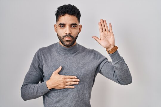Hispanic man with beard standing over white background swearing with hand on chest and open palm, making a loyalty promise oath