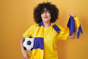 Young brunette woman with curly hair football hooligan holding ball smiling looking to the side and...