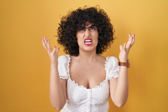Young brunette woman with curly hair standing over yellow background crazy and mad shouting and yelling with aggressive expression and arms raised. frustration concept.