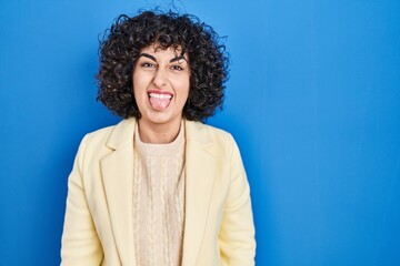 Fototapeta na wymiar Young brunette woman with curly hair standing over blue background sticking tongue out happy with funny expression. emotion concept.