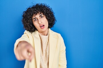 Young brunette woman with curly hair standing over blue background pointing displeased and frustrated to the camera, angry and furious with you