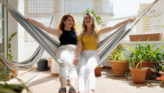 Two women smiling confident sitting on hammock at home terrace