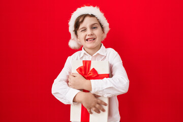 Little caucasian boy wearing christmas hat and holding gifts winking looking at the camera with...