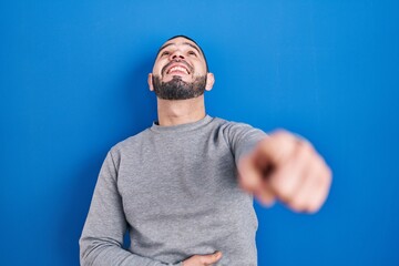 Hispanic man standing over blue background laughing at you, pointing finger to the camera with hand over body, shame expression