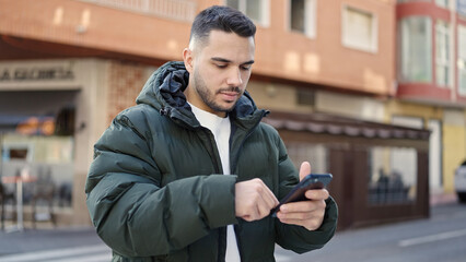 Young hispanic man using smartphone with serious expression at coffee shop terrace