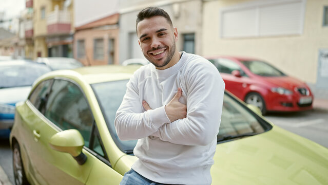 Young hispanic man smiling confident sitting on car with arms crossed gesture at street