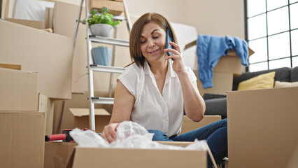 Middle age hispanic woman unpacking cardboard box speaking on the phone at new home