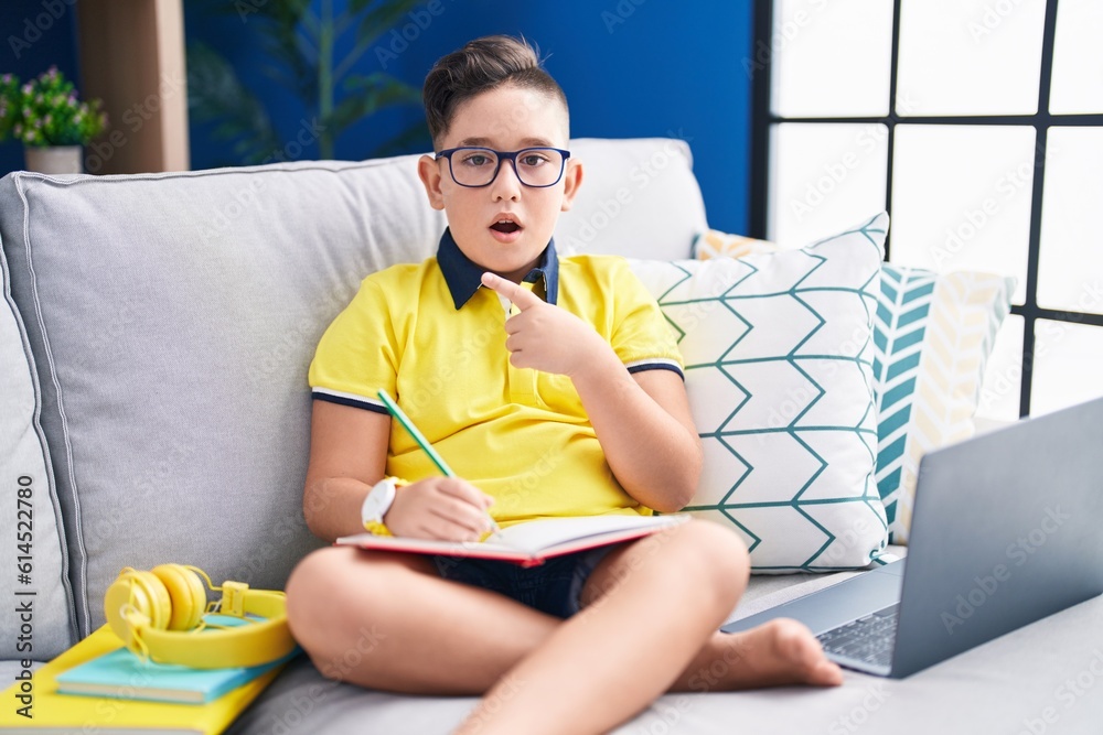 Sticker young hispanic kid doing homework sitting on the sofa surprised pointing with finger to the side, op